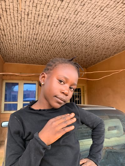 A young girl with dark skin and braided hair is in a portrait, looking directly at the camera. She is wearing a black long-sleeved shirt and a gold hoop earring. Her right hand is raised to her face. The background consists of a brick wall on the left and a window with blue panes to the right. A car is partially visible to the right of the girl. The scene appears to be outdoors, possibly in a residential area of Russo, Nigeria, during daylight hours. The lighting suggests it is daytime. There is no visible text in the image.
