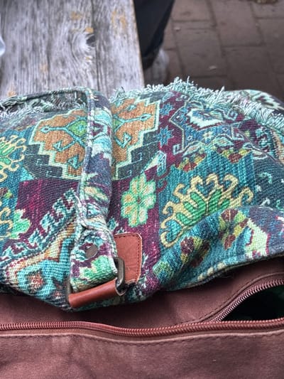 A close-up, high-angle shot shows a portion of a bohemian-style fabric bag with a fringed edge. The bag is intricately patterned with geometric and floral motifs in shades of teal, green, burgundy, and orange, resembling a Persian rug. A brown leather strap with a metal buckle is attached to the bag. Below the patterned fabric is the zipper of a brown canvas bag, which is partially open, revealing a dark interior. The bag appears to be resting on a weathered wooden surface, possibly a bench or table, with the background showing a blurred outdoor setting with cobblestone or brick paving and a person's legs and shoes.
This image was captured in Karlsruhe, Germany.
