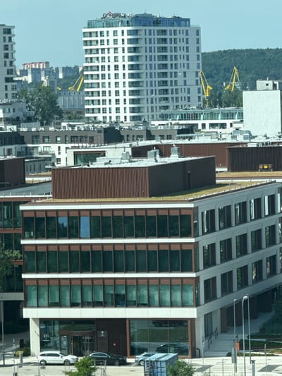 This is a medium-angle aerial photograph of a modern urban development, likely a commercial or residential complex, situated in Gdynia, Poland. The scene is dominated by several multi-story buildings with contemporary architectural designs. In the background, a tall, white, high-rise building with a blue-tinted glass facade stands prominently, featuring a logo that appears to be "CORSE". To the left of this building, another smaller white high-rise is visible. In the mid-ground, several lower-rise buildings with dark brown, corrugated metal siding are arranged in a cluster, some featuring green rooftops, indicating a possible green infrastructure element. The foreground is occupied by a multi-story building with a light-colored facade and extensive dark-tinted glass windows, framed by brown accents. This building has a stepped, angular design. At the base of this foreground building, a recessed entrance area with a revolving door is visible. Outside the building, a white SUV and a dark sedan are parked on the street. The environment appears to be a clear day with a light blue sky. The overall impression is one of a developing urban area with modern construction.