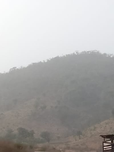 The image depicts a hazy, mountainous landscape in Jibam, Nigeria. A prominent, vegetated hill slope dominates the midground, rising towards a lighter, overcast sky that occupies the upper portion of the frame. The hill's ridge is sparsely dotted with small trees and shrubs, silhouetted against the diffused light. The lower and mid-slopes of the hill are covered in dry, brownish-green vegetation and exposed earth, with further receding hills obscured by atmospheric haze. In the bottom right foreground, a partial view of a rustic, man-made structure is visible, featuring a dark roofline and wooden support beams. No individuals, actions, or interactions are discernible.