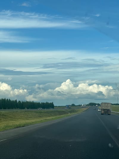 A vertical, wide-angle shot captures a highway scene under a partly cloudy sky. The image is oriented sideways, with the sky occupying the left two-thirds of the frame and the highway and its surroundings on the right. Wispy, light-colored clouds streak across a pale blue sky, with larger, more defined white clouds clustered towards the center.
On the right side of the frame, a dark asphalt highway curves gently into the distance. Several vehicles are visible on the road, including a large truck with a white container at the forefront of the highway lane. Further down the road, other vehicles, including cars and trucks, are scattered. A grassy median with a guardrail separates the highway lanes. Beyond the median, a dense line of dark green trees forms a treeline along the edge of the road. The overall scene suggests daytime travel on a rural highway. There are no visible text elements in the image.
