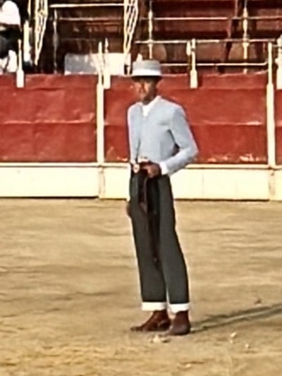 A medium shot shows a man standing in a bullfighting ring in San José del Valle, Spain. He is dressed in a light blue, long-sleeved shirt and dark pants that are rolled up to his calves, exposing white socks. The man also wears a light-colored hat and brown shoes. He holds a dark cloth draped over his left arm. In the background, there is a red wall with a white stripe at the bottom. A metal fence and seating are visible above the wall. The ground is a sandy color.