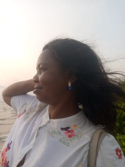 A dark-skinned woman stands in profile. Her dark hair is blowing in the wind. She wears a white button-down shirt embroidered with colorful floral designs. Her left arm is raised behind her head. A blue drop earring is visible. In the background, the sky is light, likely from the sun. Water is visible in the left background, suggesting a lakeside or seaside setting.