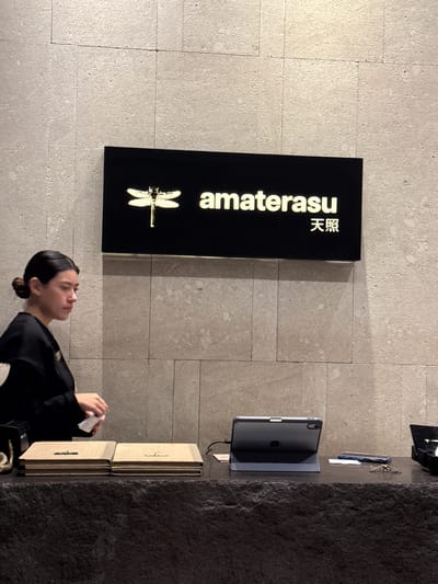 Here is a description of the image:
The image depicts an individual at a reception area in what appears to be a business establishment, potentially a restaurant, given the menus. The wall behind the person features a sign with "amaterasu" and 天照, alongside a dragonfly symbol. An iPad and other items are visible on a counter. The location is presumed to be San Pedro Garza García, Mexico.