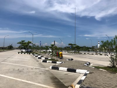 The image depicts an outdoor parking lot with a clear blue sky and scattered clouds. The parking lot is paved with concrete and marked with white lines indicating parking spaces. Several cars are visible, parked in designated spots. Along the edges of the parking lot, there are landscaped areas with trees and grass, bordered by black and white striped curbs. Streetlights are positioned throughout the parking area. In the background, there are buildings, a tall tower, and a lighthouse-like structure. The scene appears to be in Gohor Lama, Indonesia, based on the provided context, and shows a public or commercial parking facility. There are no people visible in the image.