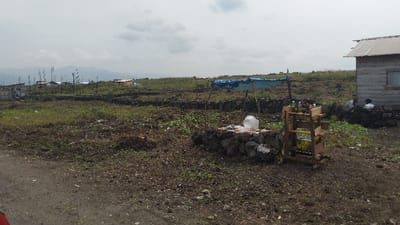 A rural landscape shows a sparse field with low stone walls and scattered buildings. In the foreground, a makeshift wooden stand holds bottles and jars next to a stone enclosure with a large plastic container. Two figures are visible near a weathered building on the right. Mountains are visible in the distance under a cloudy sky. The foreground is earthy, with patches of green and brown.