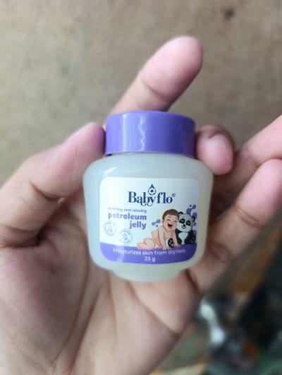 A small, clear plastic jar with a purple lid containing petroleum jelly is held in a person's hand. The jar has a white label with purple text and graphics. The label reads "Babyflo", "soothing and relaxing petroleum jelly", and "Moisturizes skin from dryness 25 g". There are cartoon illustrations of a baby and a panda on the label. The background is blurred and appears to be an outdoor setting. The person's hand is visible, with fingers curled around the jar. The location is identified as Kawit, Philippines.