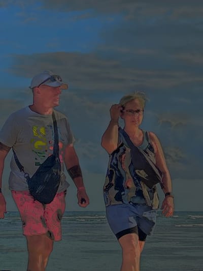 Two individuals stand on a beach, possibly in Sainte-Anne, France. One person wears a patterned shirt and shorts, along with a black Nike messenger bag. The other wears a white shirt with graphics and pink shorts, also with a black messenger bag. The background shows a cloudy sky and the ocean. The image orientation is rotated.