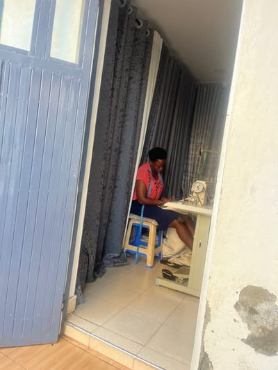 A woman sits at a sewing machine surrounded by stacks of gray curtains. She is wearing a pink top and blue skirt with a measuring tape around her neck. The sewing machine is positioned on a light-colored table. A blue and beige plastic chair is where she sits. The scene appears to be an outdoor workspace, possibly a shop entrance, as evidenced by a gray corrugated metal gate or door visible in the lower part of the frame. The lighting suggests it is daytime.