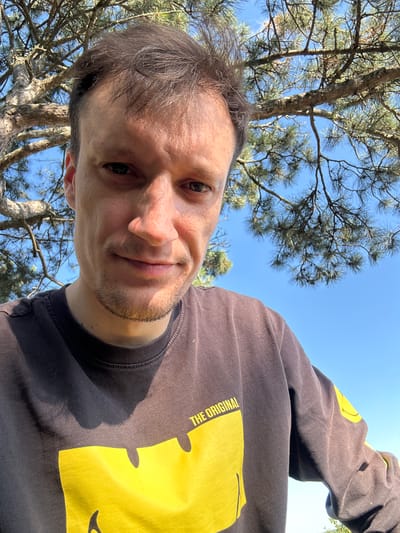 This is a selfie taken outdoors in Velykozalissia, Ukraine. A man with short, dark hair is looking directly at the camera. He is wearing a dark-colored long-sleeved t-shirt with a large yellow graphic on the front that says "THE ORIGINAL". Part of a tree with green pine needles and brown branches is visible in the upper left portion of the image, as is a clear blue sky. The man appears to be in his late twenties or early thirties, with fair skin and some stubble on his chin. The lighting suggests it is daytime, possibly late morning or early afternoon, with the sun casting shadows on the man's t-shirt.