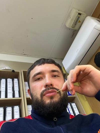 A man with a dark beard and short dark hair is in the foreground, holding a toothpick to his lips. He is wearing a dark blue fleece jacket with red trim. Behind him, shelves are filled with binders. A white air conditioning unit is mounted on the ceiling, and a white electrical box is to its left. The scene appears to be an indoor office or workspace. The image is a selfie, taken from a low angle, looking up at the man. There are a few small dark spots on the ceiling. The lighting is natural, coming from overhead.