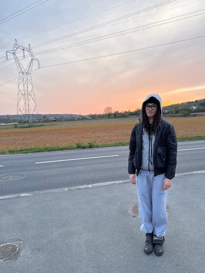 A male individual stands on an asphalt road in Marcoussis, France, facing forward. He is wearing a black beanie, rectangular glasses, a grey hooded jacket with the hood raised, a black bomber jacket, light grey sweatpants with blue lettering on the left thigh, and dark boots. The man has visible dark dreadlocks.

The scene is illuminated by twilight, with the sky transitioning from light blue to orange-pink at the horizon. To the left, a large metal lattice power pylon with multiple overhead power lines is prominently featured. The road includes white lane markings and two circular manhole covers. Adjacent to the road is a narrow grassy verge containing minimal vegetation, including a single red flower and two pink flowers. Beyond the verge, a large field of reddish-brown soil extends towards a distant treeline and hills, where faint structures are visible on the horizon.
