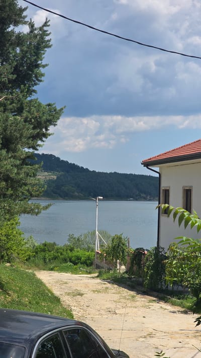 The image shows a view towards a body of water, likely the Danube River, from a property in Asparuhovo, Bulgaria. A dark-colored car is partially visible in the foreground on the left, with its roof and passenger side window showing reflections. A dirt and gravel path leads away from the viewer towards the water. On the right side of the path is the side of a white building with a red tile roof. Two windows are visible on the side of the building. Behind the building, a brick wall and green trees are present.
Beyond the path and the building, a large expanse of water stretches into the distance. Across the water, a densely forested hill or mountain range forms the horizon. The sky above is partly cloudy with a mix of dark and light gray clouds, indicating a somewhat overcast day. A single power line is visible diagonally across the upper portion of the image. A tall, white pole with a light fixture is positioned near the water's edge. Green trees and vegetation line the banks of the water and surround the path.