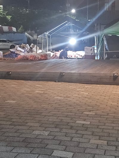 A street scene at night in Eumnae-ri, South Korea, shows a person sitting on the ground amongst a large pile of produce, likely onions, in front of a temporary tent structure. The ground is paved with brick-like stones, and there is a raised concrete platform in the foreground. Several cardboard boxes are stacked nearby, and a white car is visible in the background. The lighting suggests a market or event taking place after dark, with bright lights illuminating the area.