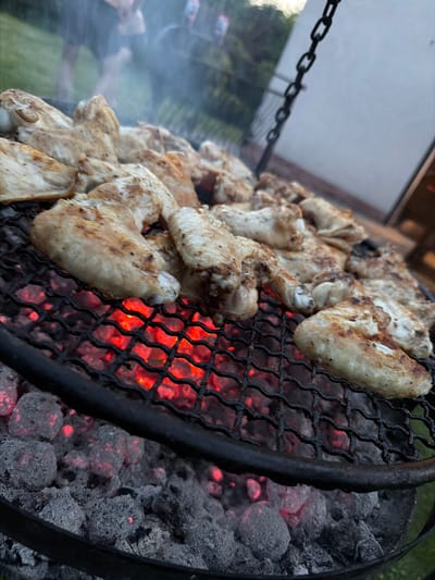 The image shows chicken wings being grilled over glowing red coals on a black metal grate. The wings are seasoned and appear to be partially cooked, with some browning and grill marks visible. Smoke rises from the grill, indicating active cooking. In the background, out of focus, are people standing in a grassy area, and a building wall. The scene is set outdoors, likely in a backyard or park, and the lighting suggests it is either late afternoon or early evening. The location is identified as Mordarka, Poland, though no specific landmarks of Mordarka are visible in the image.