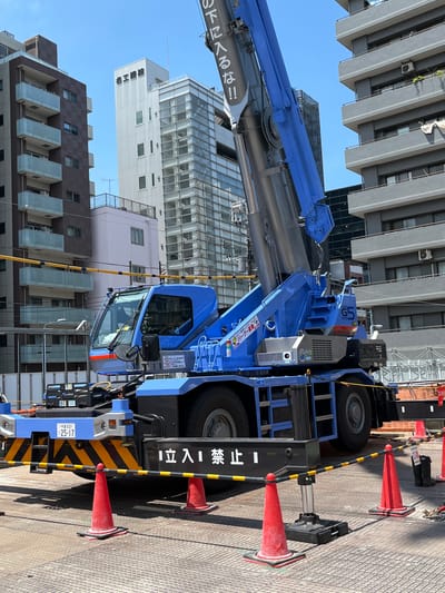 A blue mobile crane is parked on a textured ground surface in an urban environment. The crane is the primary subject, with its large boom extended upwards and to the right. The cab of the crane is visible, with tinted windows. The crane has various labels and markings, including a license plate that reads "千葉 930" and "25-17". In front of the crane, there is a black and yellow striped barrier with red traffic cones placed at intervals. In the background, there are multi-story buildings, characteristic of a city setting. The sky is clear and blue. The scene appears to be a construction or industrial site within the city of Taito, Japan. There are no people clearly visible in the image.