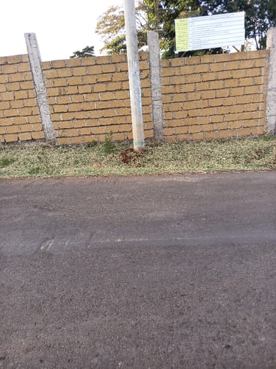 The image depicts an outdoor scene in Ewuaso Oonkidong'i ward, Kenya, during daylight hours, captured from a low vantage point looking towards a perimeter wall. In the foreground, the lower half of the image features a dark grey asphalt road surface, exhibiting a granular texture with subtle tonal variations and indistinct light markings. Directly behind the road, a narrow strip of ground consists of sparse, light green grass and exposed soil. A light grey, cylindrical utility pole stands vertically within this strip, slightly left of the image's center, with a small accumulation of reddish-brown loose soil around its base.
Behind the utility pole, a perimeter wall extends across the midground. The wall is constructed from rectangular, yellowish-brown bricks laid in horizontal courses with visible mortar joints, supported by three visible grey concrete vertical pillars. The utility pole is situated between two of these pillars.
Mounted atop the right section of the brick wall is a rectangular white signboard with dark text. The signboard details project information, including "PROJECT: RESIDENTIAL DEVELOPMENT ESTATE," "ARCHITECT: MBOI, KILAMBO & PARTNERS," "CONSULTANT: GEO-TECH CONSULTANCY LTD," and "CONTRACTOR: MEDICS SAFETY ALLIES LIMITED." Dates for "DATE OF APPROVAL" and "PLANT APPROVAL" are also visible.
In the background, above the perimeter wall, mature trees with green foliage are visible, suggesting a vegetated area beyond the wall, under a bright sky. No individuals, vehicles, or active interactions are present within the frame. The scene is static, illuminated by natural light.