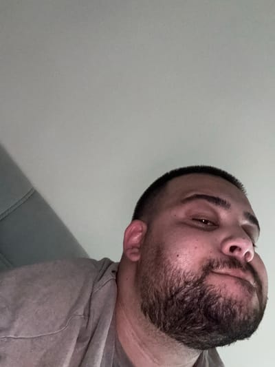 The image is a close-up, low-angle selfie of a man. The man has a short buzz cut, a full beard, and appears to be wearing a gray t-shirt. He is looking slightly to his right. The background consists of a gray ceiling and a portion of a gray upholstered surface, possibly a couch or chair, on the left side of the frame. The lighting appears to be overhead and somewhat diffuse. The location is Byala Reka, Bulgaria.