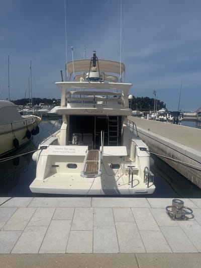 The image shows a yacht named "Aurora," available for rent (www.sixjyacht.com), docked at a marina in Grad Rovinj, Croatia. The yacht is white with a multi-tiered structure, a ladder leading to the upper levels, and "Yacht for rent" printed on its stern. It is docked alongside a stone quay with metal mooring fixtures. Other boats and buildings are visible in the background. The sky is blue and clear.