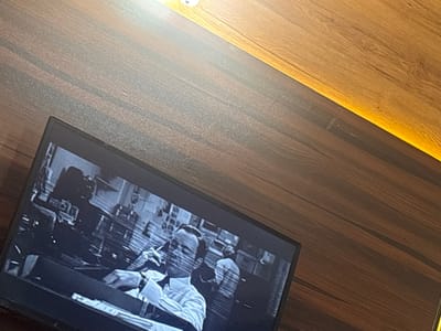 A television displays a black and white film scene with a man seated at a desk, wearing a tie and collared shirt. He is illuminated by a desk lamp and appears to be smoking. Behind him, another figure is visible, also in a collared shirt. The television is placed on a wooden surface. There is no discernible indication of the location being Eneka, Nigeria.