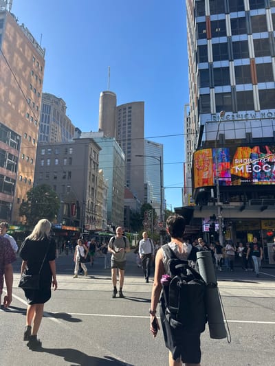 A street scene with pedestrians walking along the asphalt. Buildings of varying heights line the street under a clear blue sky. Among the shops is a "Mecca" sign. The people are walking in a crosswalk and the street is oriented vertically.