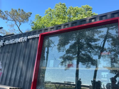 The image shows a close-up of a coffee shop with a cracked glass window. The coffee shop appears to be constructed from a shipping container, painted dark gray with corrugated siding. White lettering on the side of the container reads "ALAGAMTA COFFEE". The large window in the foreground is framed with red metal and is heavily cracked, with the glassweb-like pattern of the damage visible.
Through the cracked window, reflections of trees and a clear blue sky can be seen. Inside the shop, some objects are visible, including shelves and what appear to be tables and chairs. A red Coca-Cola logo is visible as a reflection on the glass. The overall scene suggests an outdoor setting on a sunny day.
The time of day appears to be morning or afternoon, given the bright sunlight and clear sky. The presence of lush green trees indicates a warm season, likely spring or summer. The broken window suggests an incident may have occurred, potentially causing a sense of disruption or disrepair, though the bright weather and the presence of the business suggest it may still be operational or in the process of repair. The location context of Ochamchyra, Abkhazia, is noted.
