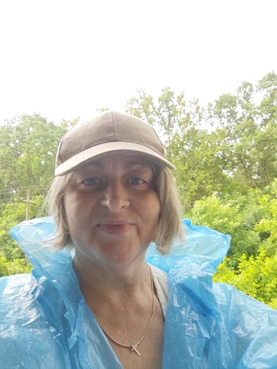 An outdoor, waist-up, eye-level selfie shows a middle-aged woman with shoulder-length blonde hair, wearing a tan baseball cap and a bright blue disposable poncho. She is looking directly at the camera with a neutral expression. Her eyes are blue, and she has light skin. A thin silver necklace with a cross pendant is visible around her neck. Behind her, the blurred background consists of lush green trees and foliage, with a glimpse of utility poles and wires on the left, suggesting an outdoor, possibly rural or park-like setting in Znamenskiy, Russia. The sky is overcast and bright white. The poncho appears slightly damp, and there are visible wrinkles and folds in the plastic material.