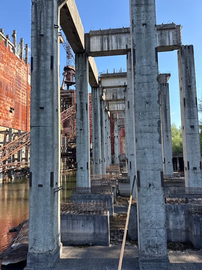 The photograph depicts the skeletal remains of a large industrial complex under a clear blue sky. Massive, weathered concrete pillars and beams dominate the foreground, some showing signs of decay with exposed rebar and chipped surfaces. These structures are arranged in a regular, repetitive pattern, suggesting a former assembly line or processing area.

In the mid-ground, rusted metal structures, including what appears to be a dismantled crane and a large, reddish-orange industrial wall, rise above the murky water. The water reflects the sky and the surrounding concrete, creating a slightly distorted mirror image. The presence of exposed rebar and unfinished concrete foundations indicates that the construction or deconstruction of this site was either halted or is ongoing.

The environment appears to be an abandoned or decaying industrial zone, possibly on the outskirts of a city. The lack of human presence and the state of disrepair suggest neglect and the passage of time. The overall impression is one of industrial grandeur reduced to ruins, with nature beginning to reclaim the man-made structures.

While the prompt mentions Kyiv, Ukraine, this specific location is not explicitly identifiable from the visual elements alone. The scene is characterized by industrial decay, concrete structures, rusted metal, and water.