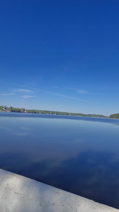 The image presents a view of a lake or wide river under a clear blue sky. A thin strip of land with vegetation and some structures lines the opposite shore. A white, possibly concrete, barrier or railing is visible in the foreground on the right side of the image. The image is rotated ninety degrees counterclockwise, making the sky appear on the left and the water vertical. There is no information to place the location in the city of Otchor, Russia.
