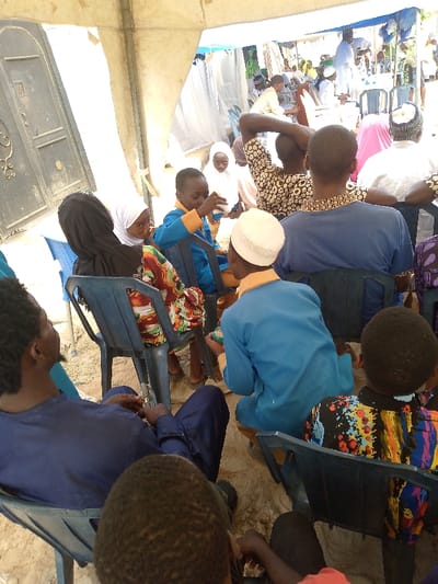 A gathering under a tent shows people seated in plastic chairs. Many are in traditional Islamic attire, including headscarves and kufis. A child is drinking from a small cup. A variety of colorful clothing patterns are visible. A closed gate is on the left. The setting appears to be outdoors, with sand visible on the ground. The atmosphere seems relaxed and communal.