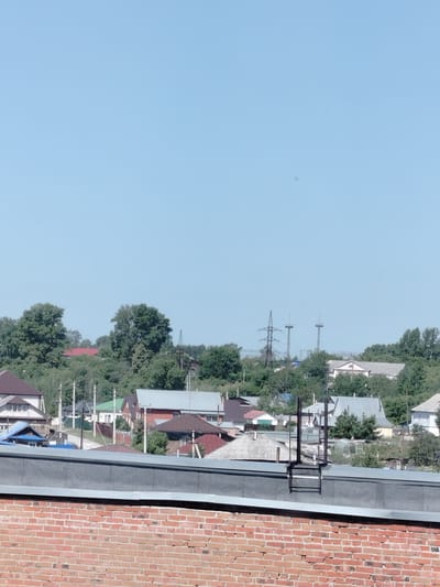 The image depicts a clear, blue sky over a residential area in Yashkino, Russia. In the foreground, a brick wall forms the bottom of the frame. Above the wall, a flat roof with a metallic edge and a dark grey surface is visible, featuring a metal ladder structure. Beyond the roof, a collection of houses with various roof types, including peaked and flat roofs, are nestled amongst lush green trees. Some houses have red, brown, and grey roofs. In the background, a large electrical pylon and two tall, slender towers with elevated platforms are prominent against the skyline, indicating the presence of infrastructure. The overall scene suggests a sunny day in a suburban or rural setting.