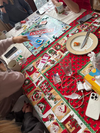The image displays an overhead view of a table in an indoor setting located in Нахабино Кантри Кп, Russia. The table is covered by a red and green Christmas-themed tablecloth featuring patterned squares with Santa Claus figures, Christmas trees, snowmen, and stockings.
At least four individuals are present, evidenced by visible hands, arms, and torsos positioned around the table. In the upper left quadrant, a person wearing a dark grey long-sleeved top is partially visible, with a silver-colored calculator and a small red rectangular object on the white section of the tablecloth. Directly in front of this area is a Russian version of the Monopoly board game, titled "МОНОПОЛИЯ," with green house/hotel tokens, player pieces, and blue property cards arranged on various spaces. A person's hand, wearing a light-colored sleeve, is seen pointing towards the game board.
In the upper right quadrant, a person in a red top holds a white mug with a festive character design. A silver mobile phone with multiple cameras rests on the tablecloth nearby. To the right, a round, light-colored plate with a gold rim holds a single rectangular food item topped with orange roe and a silver knife. A crumpled white tissue and an empty clear glass are adjacent.
In the lower right quadrant, a yellow and blue "TOLLI" brand tissue box, a white mobile phone charging plug, a white charging cable, a white wireless earbud case, and a white mobile phone housed in a patterned white case with multiple cameras are displayed. A blue and white Monopoly card lies next to another clear empty glass.
In the lower left quadrant, a person with blonde hair is partially visible, with their hands clasped. A white mug containing a light brown liquid, a stack of Monopoly property cards, and a black mobile phone are situated on the tablecloth. The overall scene indicates an active board game session with accompanying snacks and beverages.
