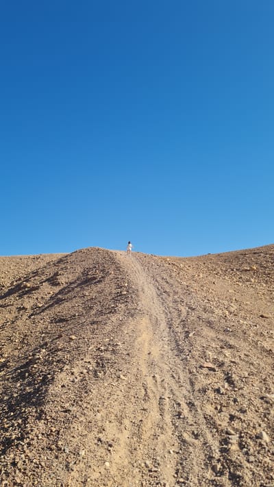 A person stands on the edge of a rocky, sandy cliff under a clear blue sky. The ground is covered in loose rocks and sandy soil. The image appears to be taken in Eilat, Israel, judging by the arid landscape. The person is small in relation to the environment, emphasizing the scale of the cliff and the open sky.