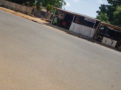 This media file shows a view of a road in Palade, Nigeria, with a small roadside restaurant on the right. The road is paved and appears to be in moderate condition. The restaurant has a corrugated metal roof supported by wooden beams, and its walls are made of vertical wooden slats painted white. There is a sign above the entrance that reads "RESTAURANT" in a decorative font, with a logo featuring a musical instrument. A satellite dish is visible on the roof of the restaurant.

Two people are seated at a table outside the restaurant, one at a green plastic chair and the other at an orange plastic chair. Another person is standing behind the counter inside the restaurant. The scene suggests a casual dining setting. In the background, there is a wall with some graffiti on it, which appears to say "DOKA TAHAN FITSARI" and other illegible text. A bamboo fence runs along the top of the wall. Trees are visible in the background, suggesting a semi-rural or suburban environment. The weather appears to be clear and sunny, indicating it is daytime. The overall impression is of a typical roadside establishment in a warm climate.