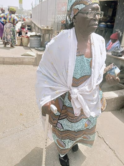 The image captures an outdoor scene in Kadoma, Zimbabwe, centered on an elderly dark-skinned woman in the foreground. She wears rectangular spectacles, a patterned headscarf, and a matching patterned dress in teal, brown, and orange hues. A white shawl, subtly speckled with gold, is draped over her shoulders and knotted at her waist. She holds a silver walking stick with a white handle in her left hand and folded currency notes in her right hand. Her feet are clad in dark open-toed sandals. Her head is turned towards the right, with her gaze directed off-frame.

In the mid-ground, a market environment is visible. To the left, two women in patterned clothing are partially visible, facing away. A vendor, identified by a red top, is seated behind multiple baskets containing red produce, likely onions. Adjacent to the vendor is a large, grey cylindrical container with a lid. Further behind, various market stalls with corrugated metal siding are present. On the right mid-ground, a person wearing a red hat is seated near an obscured stall entrance. The ground is a light-colored, paved surface, and the scene is illuminated by direct sunlight. Distant buildings and a street are discernible in the far background.