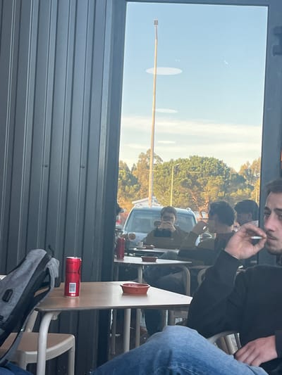The image, originally rotated 90 degrees clockwise, depicts a lively daytime scene, likely from within a café or restaurant, looking out through a large window.
In the foreground, on the right side of the frame, a young man with dark hair is visible in profile, holding a lit cigarette between his fingers. He wears a dark shirt. Below him, the lower body of another individual in blue jeans and a white shoe is partially seen. A light-colored wooden table extends from the bottom right towards the center, bearing a red ashtray and a red "COCA-COLA" can lying horizontally. A grey backpack rests below the table in the bottom left. The textured grey exterior wall of the building occupies the lower left portion of the image.
The large window offers a view of the outside, reflecting elements from inside as well. A prominent reflection shows a man wearing glasses and a light-colored top, seemingly looking down at something. Behind him, a silver car is parked. Through the window, a bright, clear sky dominates the background, with a long, slender metal light pole stretching across it. Lush green trees are visible further in the distance. The overall lighting suggests it is a sunny day.
The scene portrays a casual gathering, with individuals relaxing, and one person smoking. The multiple reflections create a layered effect, blending the interior and exterior environments. The location context is Pombal, Portugal.