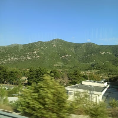 The image depicts a natural landscape with a mountainous backdrop and a clear blue sky. In the foreground, a road is visible, suggesting a view from a moving vehicle. To the right, a modern building with a flat roof and white walls is partially obscured by lush green foliage. The building appears to be a commercial or industrial structure, featuring vents and possibly an air conditioning unit. The mountains are covered in dense green vegetation, with some rocky outcrops visible. A telecommunications tower can be seen on the summit of one of the peaks. The overall scene suggests a location in a rural or semi-rural area, possibly on the outskirts of a town or city. The presence of the building and the road indicates human activity and infrastructure within this natural setting. Given the context provided, this scene is likely situated in the Municipal Unit of Ampelakia, Greece, a region known for its mountainous terrain and scenic beauty.