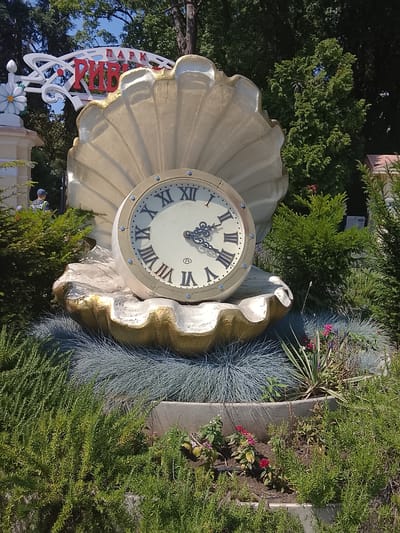 The image depicts an outdoor scene featuring a large decorative clock shaped like a clam shell, situated in a park setting. The clock has Roman numerals and ornate hands. In the background, there is a sign with Cyrillic lettering that reads "ПАРК РИВЬЕРА" (Park Riviera), indicating the location is in Oryol-Izumrud, Russia. The clock is surrounded by lush greenery and flowering plants, with trees forming a canopy overhead. The overall atmosphere is bright and sunny, suggesting a pleasant day in the park.