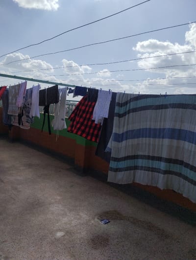 The image depicts an outdoor scene, likely a rooftop, in Witeithie ward, Kenya, on a bright day under a partly cloudy sky. Numerous garments are suspended on multiple clotheslines strung across a green metal frame. The drying laundry includes a red and black plaid shirt, various solid and patterned shirts, dark trousers, and a large striped blanket or sheet featuring blue, white, and grey/tan bands. These items are secured to the lines with blue clothespins. Below the clotheslines, a low wall or barrier is visible, painted with horizontal sections of brown and green. The ground surface is a mottled grey concrete. In the lower right foreground, a small, dark blue or purple rectangular object, possibly a wrapper or packet, rests on the ground. Overhead, several thin, dark wires traverse the blue sky, which contains scattered white cumulus clouds. No individuals are present in the scene.
