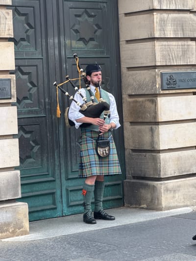 A male bagpiper stands in front of large, dark green double doors with geometric carvings. He wears a black Glengarry hat, a white shirt, a teal vest, and a kilt in a tartan pattern of green, blue, and red. He also wears green knee-high socks with orange flashes peeking out, and black shoes. He is playing the bagpipes, holding them with his right arm and blowing into the chanter with his mouth. A sporran hangs from his waist. To his right, on the stone building, is a plaque that reads "HIGH COURT". The building is constructed of large, grey stone blocks. The scene is outdoors, with a grey pavement in the foreground. This is the High Court of Justiciary on Parliament Square, City of Edinburgh, United Kingdom.