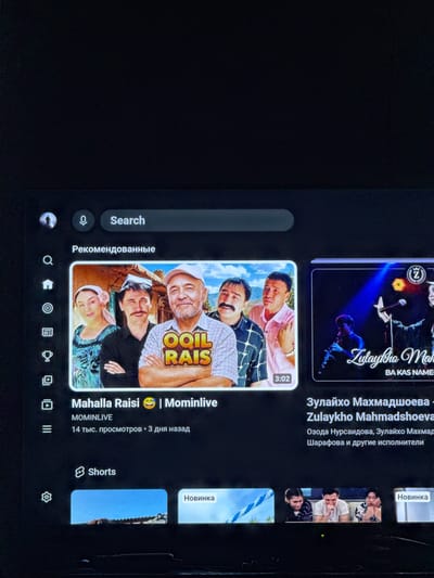 This is a media file that appears to be a screenshot of a YouTube-like interface, likely on a mobile device or tablet, given the aspect ratio and interface elements. The content displayed is a mix of video thumbnails and interface options.
The main subject is a collection of video thumbnails featuring people, predominantly of Central Asian descent, dressed in traditional attire. One prominent thumbnail shows three individuals, two men and a woman, with the text "OOIL RAIS" overlaid. Another thumbnail features a performer on a stage with vibrant lighting, accompanied by Cyrillic text that translates to "Zulaykha Mahmadshoeva". There are also thumbnails labeled "Shorts" and "Новинка" (New).
The interface elements include a search bar with a microphone icon, a "Search" label, and "Рекомендованные" (Recommended). At the bottom, there's a navigation bar with icons suggesting options like home, search, subscriptions, library, and settings.
The overall scene suggests a user browsing or interacting with video content, possibly entertainment or music. The presence of Cyrillic text and the appearance of the individuals strongly suggest a context related to Uzbekistan, and the prompt specifies Bukhara, Uzbekistan, as the location. The images evoke a sense of cultural performance and everyday life. There are no explicit indicators of time of day or weather. The emotions conveyed by the individuals in the thumbnails range from smiling to more neutral expressions.