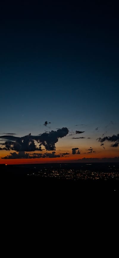A wide-angle, vertical landscape photograph captures a dusky sky above a city at dusk. The sky transitions from a deep, dark blue at the top to a fiery orange and red hue near the horizon, indicating the last moments of daylight. Dark, dramatic clouds are scattered across the sky, some appearing to be illuminated from below by the fading light.

Below the sky, the dark silhouette of a city is visible, with countless small, glowing lights representing buildings and streetlamps. A faint horizontal line suggests a distant horizon, possibly marking the edge of the city or surrounding landscape. The foreground is almost entirely black, representing the earth or a higher vantage point looking down on the scene.

The image appears to be taken from an elevated position, offering a panoramic view of the city as night begins to fall. The overall mood is serene and contemplative, with the stark contrast between the dark foreground and the illuminated city creating a sense of depth and mystery. The presence of a contrail from an aircraft is subtly visible in the upper portion of the sky, adding a touch of human activity to the otherwise natural scene. The specific location in Lyaskovets, Bulgaria, is not discernible from the image itself, but it depicts a typical urban landscape at twilight.