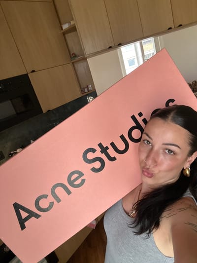 A selfie shows a woman with dark hair and fair skin, wearing a gray tank top and gold jewelry, posing with an "Acne Studios" box. The surroundings include wooden cabinets, a reflective black surface and a window showing buildings outside, indicating an interior setting in Saint-Ouen-sur-Seine, France.