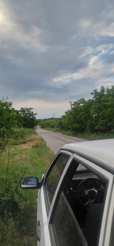 A white car is parked on the side of a gravel road in Ketrysanivka, Ukraine. The car's driver's side door is open, revealing the steering wheel and part of the interior. The road stretches into the distance, flanked by lush green trees and vegetation. Overhead, a cloudy sky with hints of blue suggests an overcast day. Power lines are visible strung between utility poles along the side of the road. The scene appears to be a rural or semi-rural area.