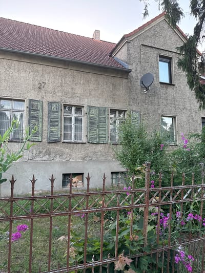 The image depicts the facade of a two-story house, likely in Wustermark, Germany, given the context. The house has a textured, light grey stucco exterior and a reddish-brown tiled roof. The number "27" is visible on the upper story. Several windows are present on the facade, some with weathered, green wooden shutters. The windows are fitted with patterned curtains. A satellite dish is mounted on the upper right side of the house. A rusty metal fence with pointed finials runs along the front of the property, separating it from a garden area with lush green foliage and small purple flowers. The garden appears somewhat overgrown. The overall impression is one of an older, perhaps slightly neglected, residential property.