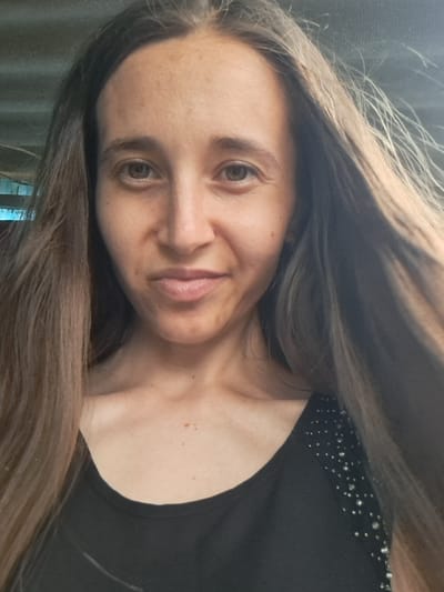 This is a close-up, rotated portrait of a woman. She has long, brown hair that falls around her face and shoulders. She is wearing a black shirt with small, shimmering embellishments along the neckline. The background is out of focus and appears to be a textured surface. The lighting suggests it is daytime, likely indoors. Her expression is neutral, and her eyes are looking towards the viewer. The image is a selfie, taken from a slightly high angle. There is no visible text in the image. The provided location context is Vypasne, Ukraine.