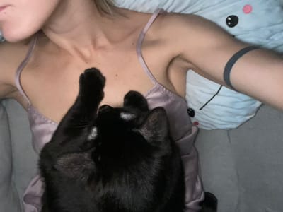 The image shows a young woman lying in bed with a black cat. The woman is wearing a pink satin top and has a tattoo on her left arm. The cat is lying on her chest with its paws near her neck. The woman's right arm is extended above her head and her left arm is lying by her side. The bed linens are gray. The image was taken in the city of Moscow, Russia at the coordinates 55.659, 37.591.