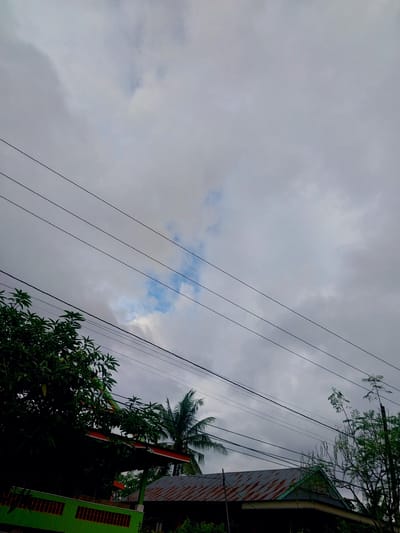 The image captures a cloudy sky over a residential area in Turumame, Indonesia. Several dark power lines crisscross the frame horizontally. In the foreground, lush green foliage from trees partially obscures a bright green fence with orange accents. Beyond the trees, the roof of a house is visible, constructed with corrugated metal, showing signs of rust. To the right, a portion of another house with a similar metal roof and a green trim is visible. A palm tree and other trees with green leaves are scattered throughout the scene. A small patch of blue sky is visible through the clouds. The overall impression is one of a typical overcast day in a tropical urban or suburban setting.