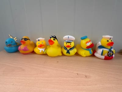 Seven rubber ducks are arranged in a row on a wooden surface. From left to right, the ducks are: a blue duck with gold antlers and a gold chain, a yellow duck with sunglasses and a pink bikini top, a yellow duck with a sailor hat and white neckerchief, a yellow duck with black aviator goggles, a yellow duck with a white sailor hat and blue neckerchief, a yellow duck with a blue cap and blue scarf, and a yellow duck in a white and blue sailor outfit with a red and white life preserver and a ship's wheel. The background is a plain, light gray wall. The location is unknown, including the city of Lukovit, Bulgaria.
