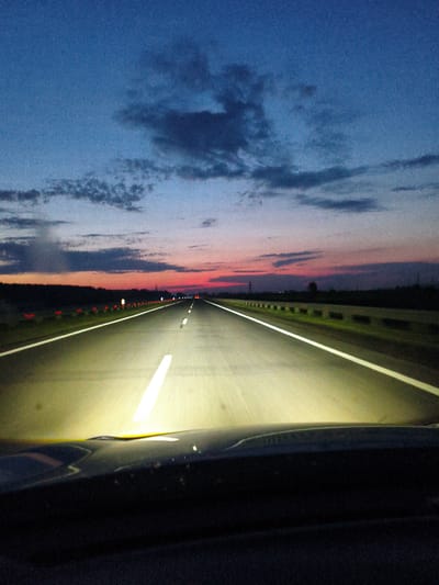 The image captures a view from a vehicle driving on a highway at dusk. The sky transitions from a deep blue at the top to hues of pink and orange near the horizon, with scattered dark clouds. The highway is illuminated by the headlights of the vehicle, casting a bright beam on the asphalt and the white lane markings. A dark silhouette of trees and distant structures lines the side of the road. The foreground shows the dashboard of the vehicle. There are no people or specific landmarks visible that would indicate the location within Polysayevo, Russia.